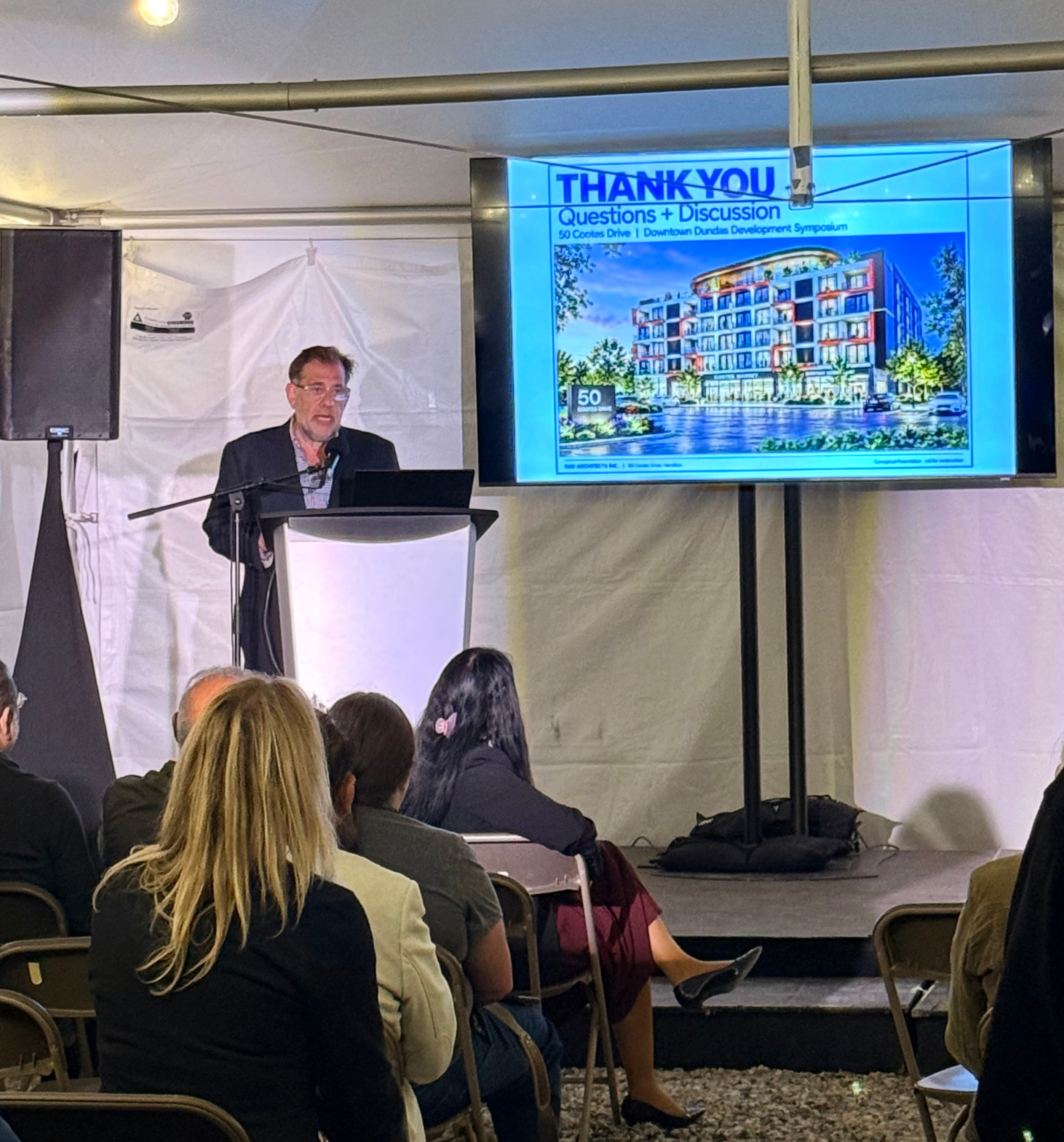 Loren Lieberman at a podium inside an event tent, a screen beside him showing a rendering of a mid-rise building at 50 Cootes Drive under the words Thank You, Questions + Discussion, with an audience seated in front