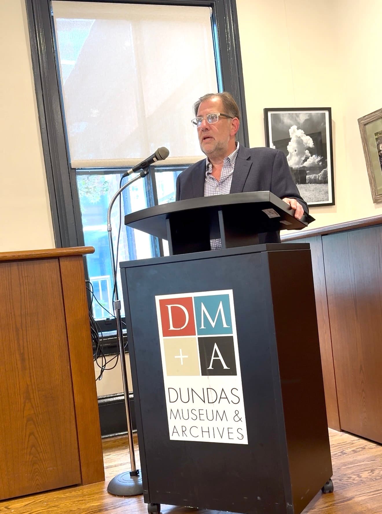 Loren Lieberman speaking into a microphone at a Dundas Museum & Archives podium, a tall window and framed photographs behind him
